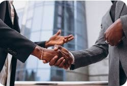 Deux personnes en tenue professionnelle se serrent la main devant un immeuble de bureaux.